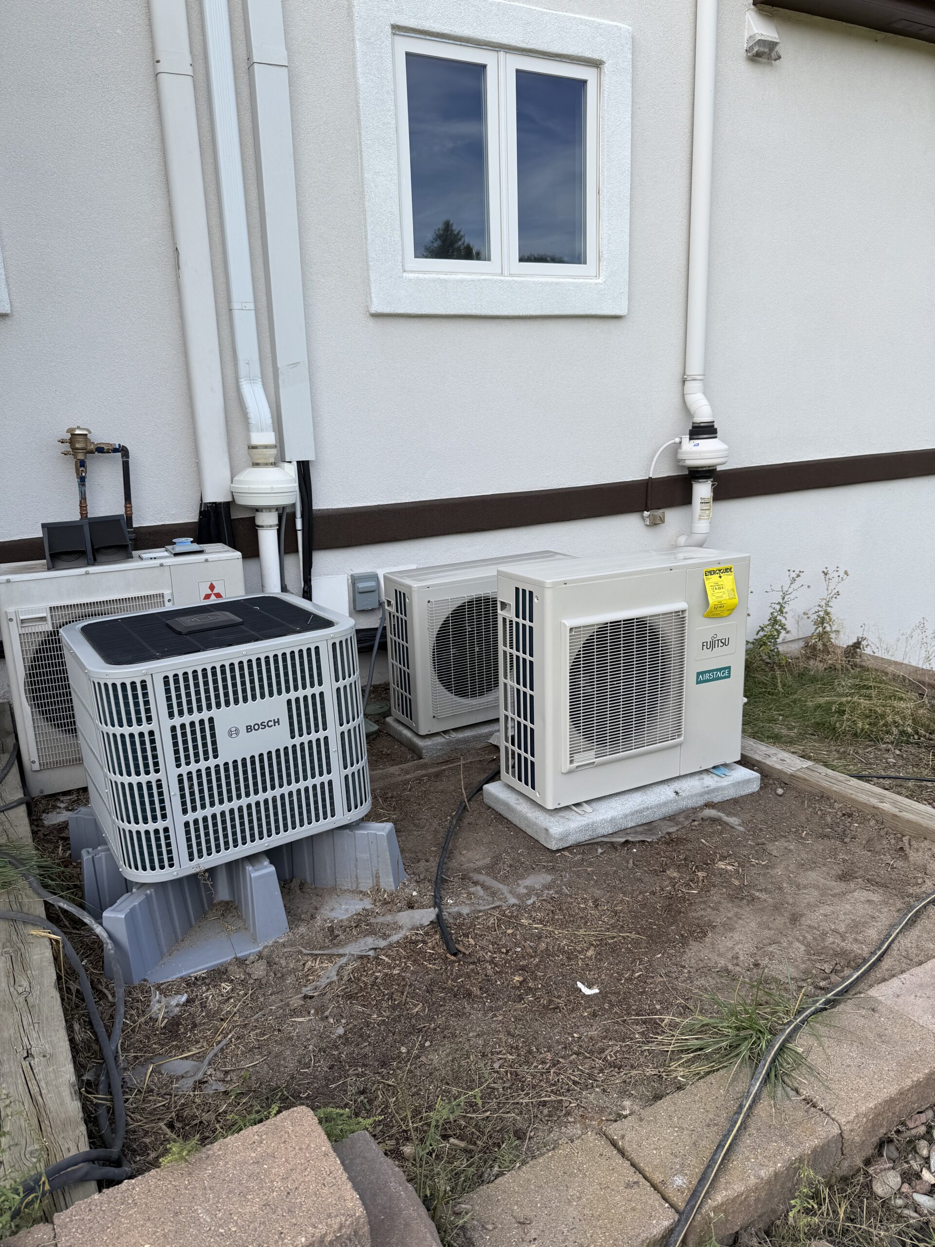 Three outdoor HVAC units are installed on concrete pads next to a white building, with cables and pipes connected to the wall. Grass and dirt are visible in the foreground.