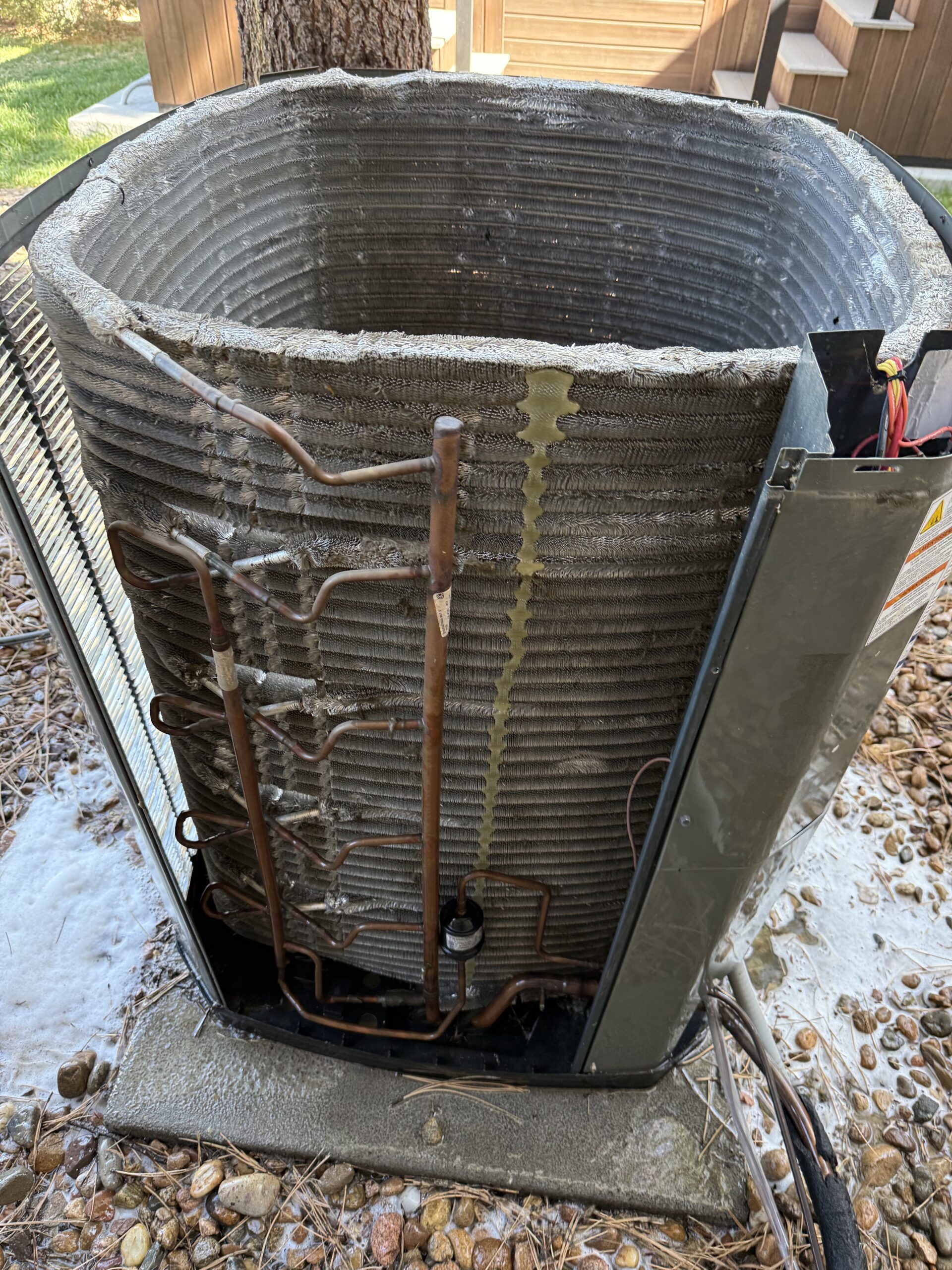An outdoor air conditioning unit with its outer cover removed, showing exposed coils and copper tubing; a yellowish liquid stain is visible on the coils.
