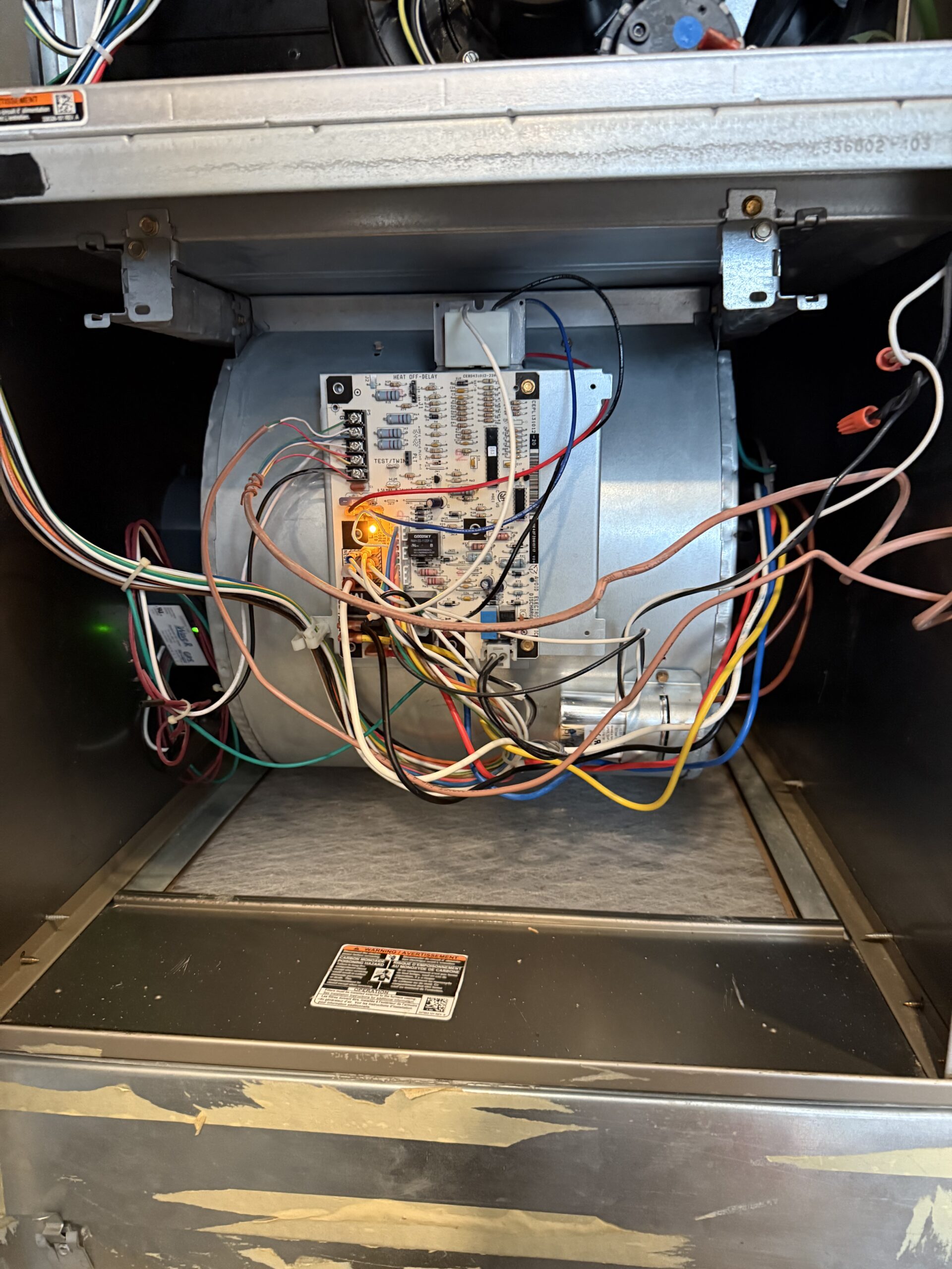 An open HVAC unit showing a circuit board with multiple colored wires connected, mounted on a silver blower assembly, with a visible air filter below.