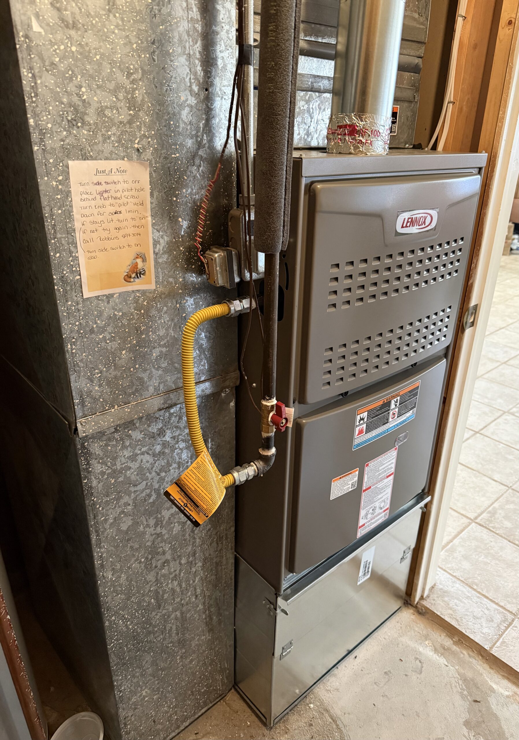 A gray Lennox furnace with attached pipes and a yellow gas line stands next to a metallic duct in a utility room; a note is taped to the duct.
