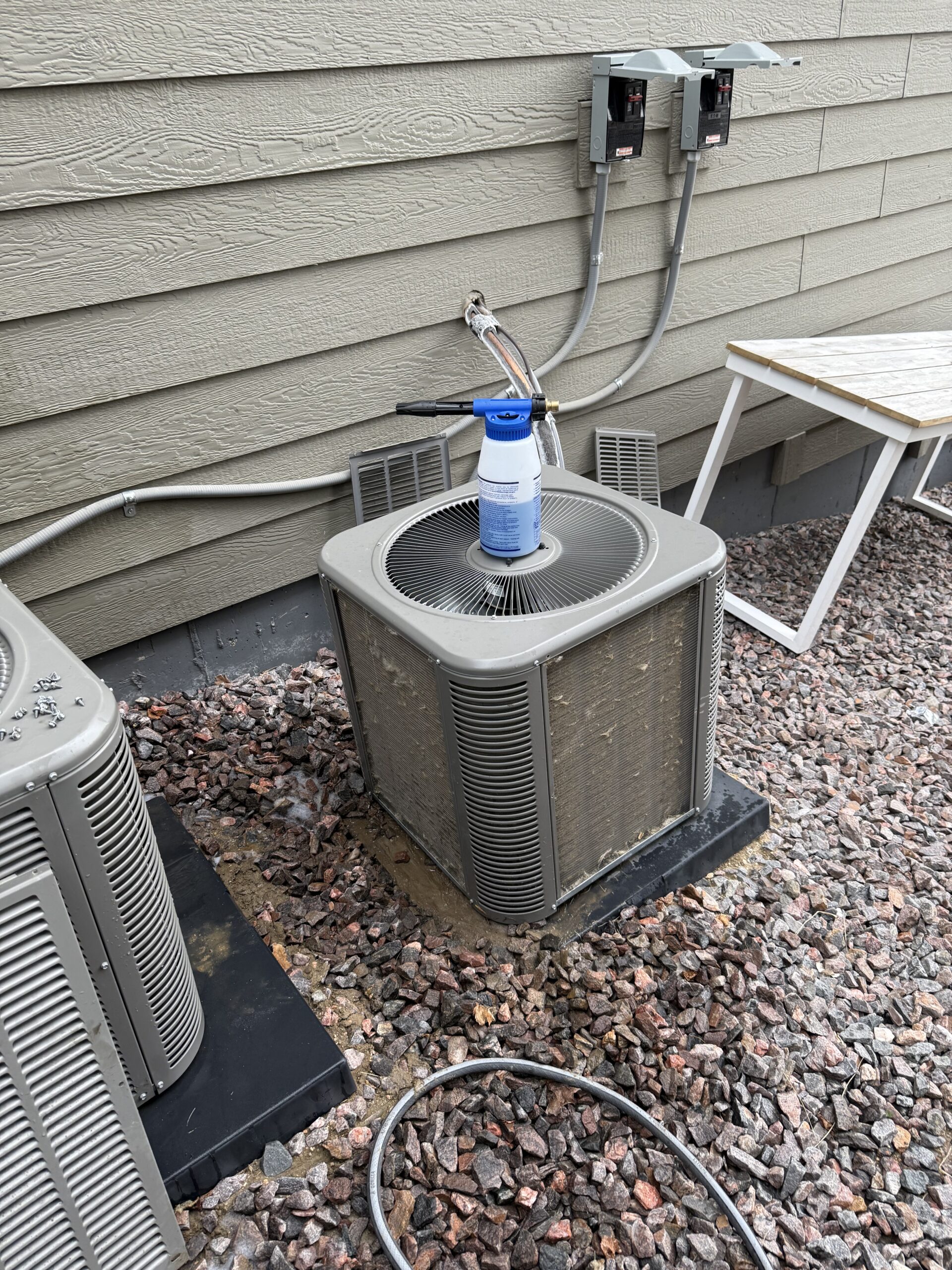 A whole house water filter rests on top of an outdoor air conditioning unit next to a house with exposed wiring and a rock-covered ground.