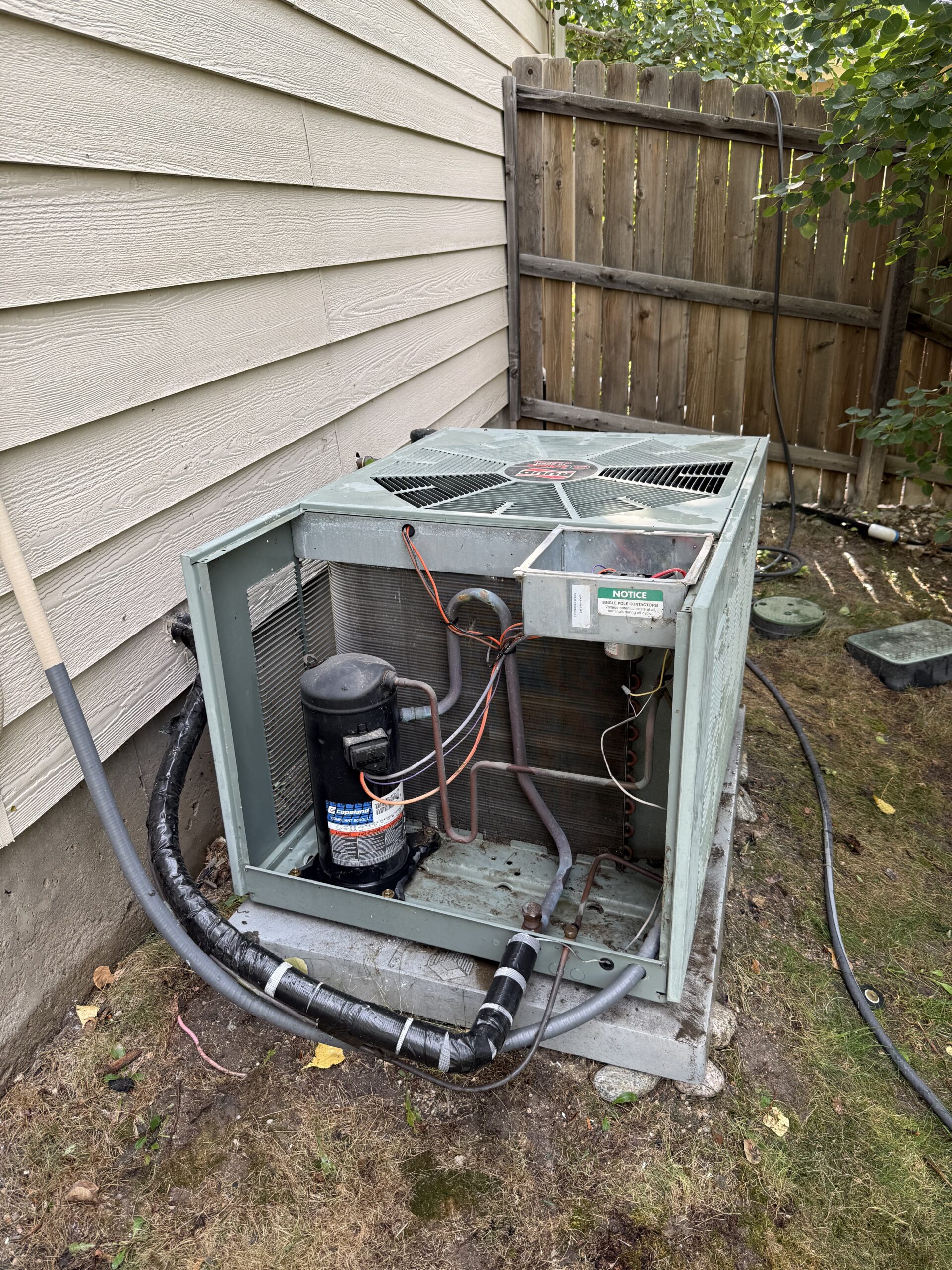 An outdoor air conditioning unit with its cover removed sits on a concrete pad next to a house, exposing internal components and wiring.