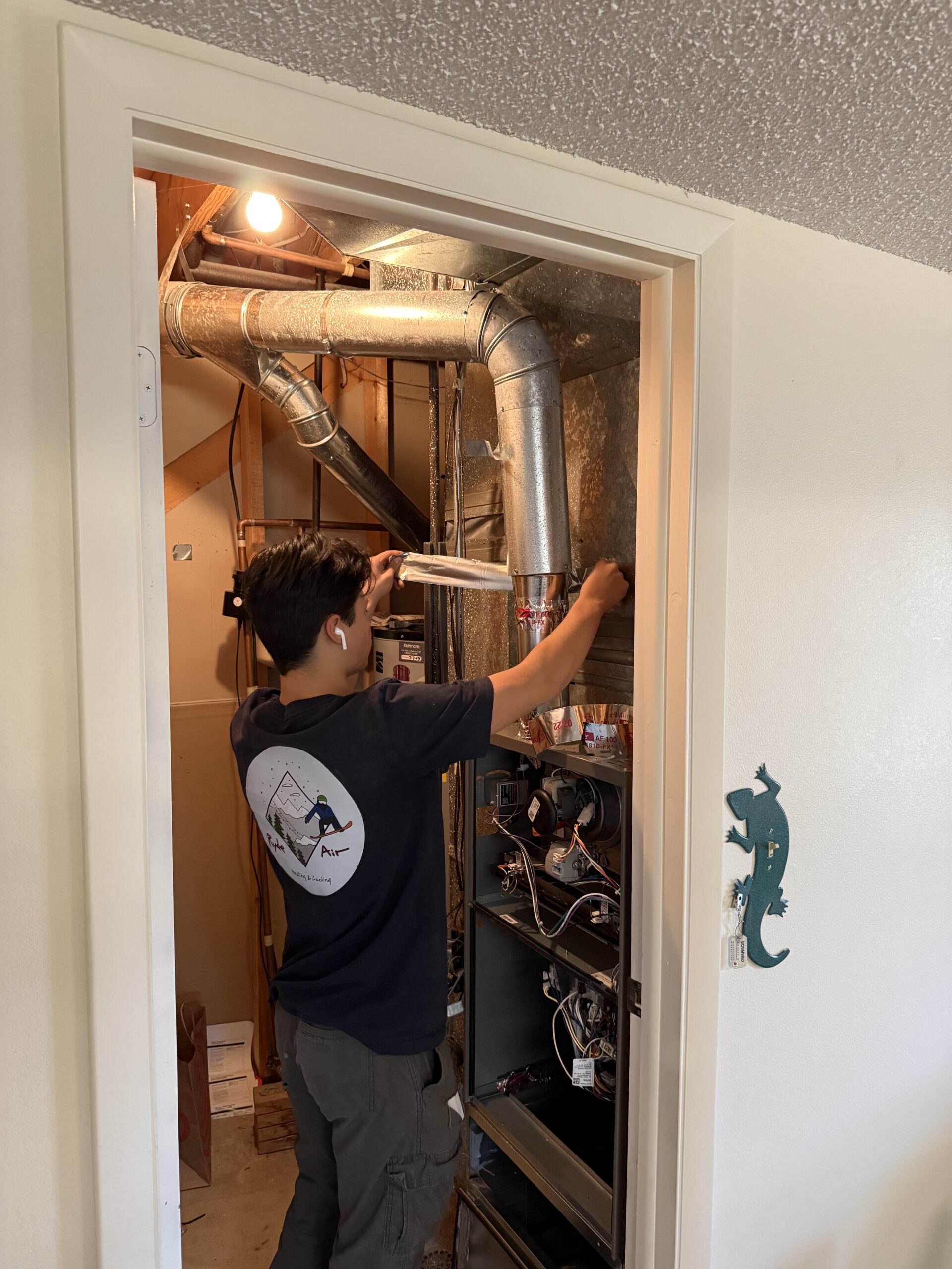 A person stands in a small utility closet inspecting or repairing an HVAC unit with tools. Ductwork and wires are visible above, and a green lizard decoration is on the door frame.