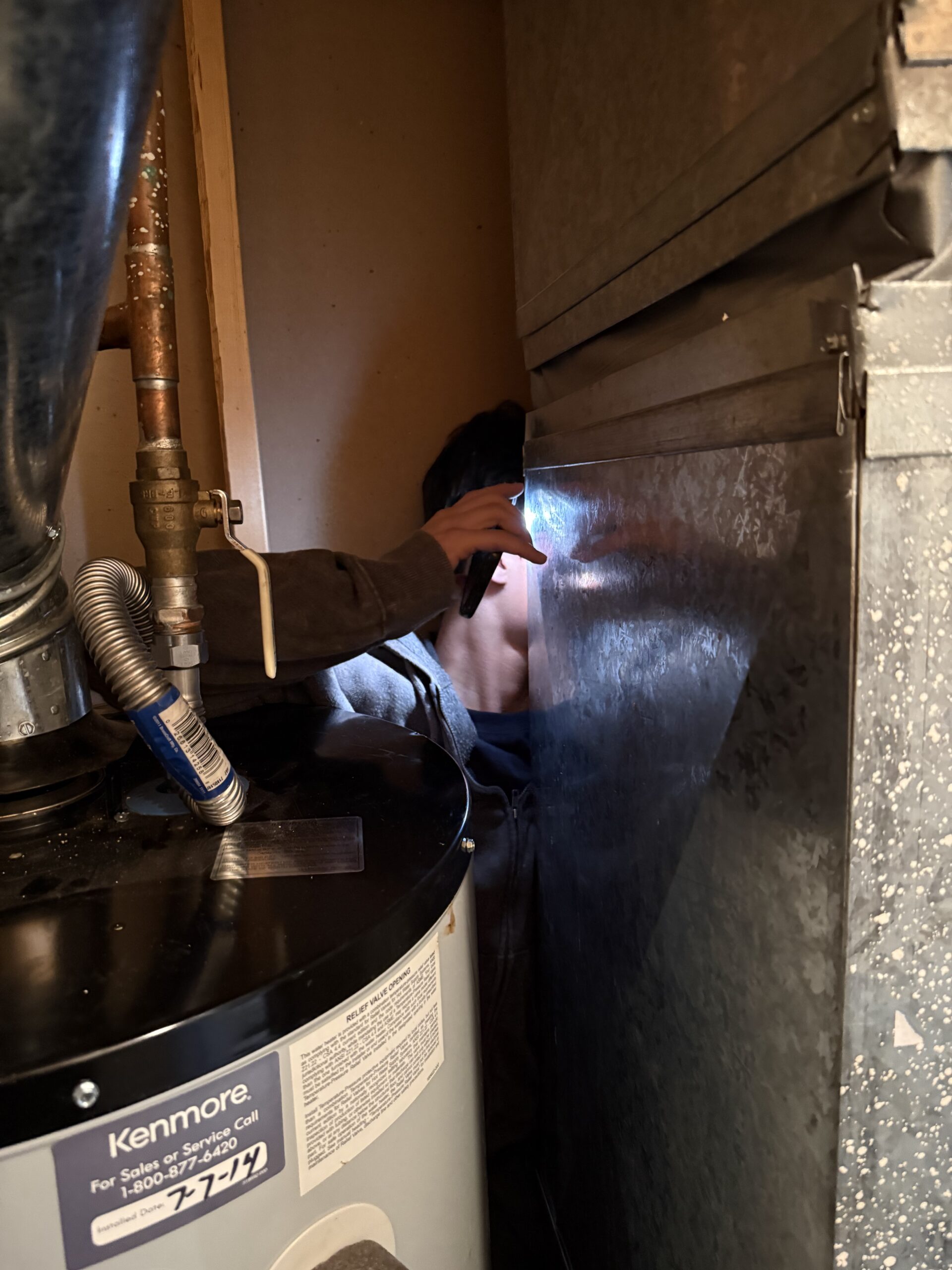 Person shining a flashlight and inspecting a metal vent or duct behind a hot water heater in a narrow utility space.