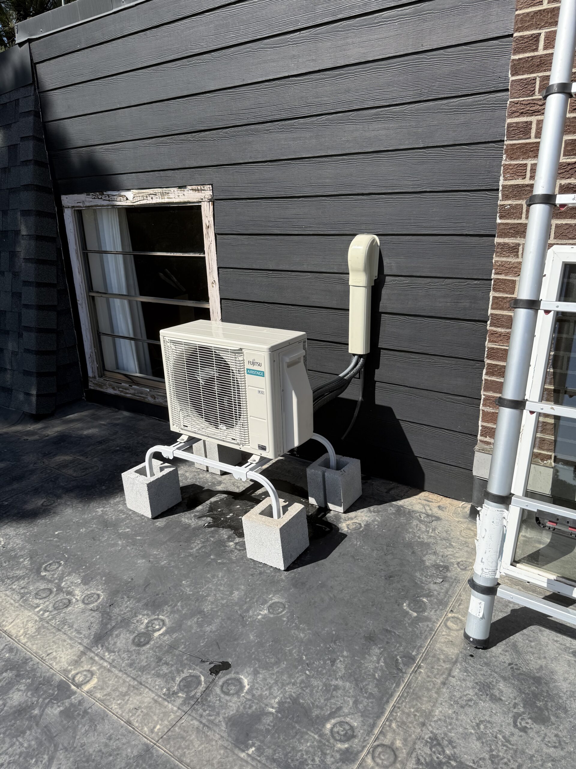 Outdoor air conditioning unit mounted on concrete blocks beside a dark-sided building, with cables connected to a vertical utility box on the wall.